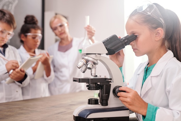 Умная серьезная школьница в белом халате смотрит в микроскоп за партой на фоне своих одноклассников и учителя, проводящих химический эксперимент
