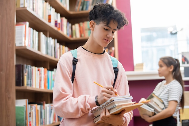 Умный студент-подросток с карандашом делает заметки в блокноте, стоя у большой книжной полки в библиотеке