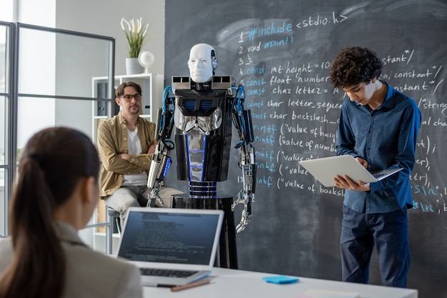 Photo clever teenage guy with laptop standing by blackboard in front of class and making presentation of robot abilities and characteristics