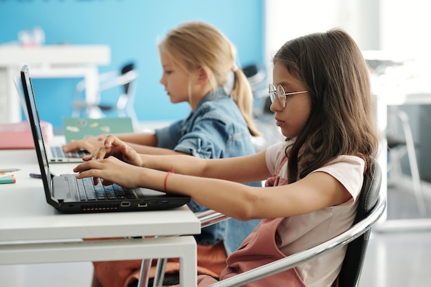Умная юная школьница и ее одноклассник печатают на клавиатуре ноутбука