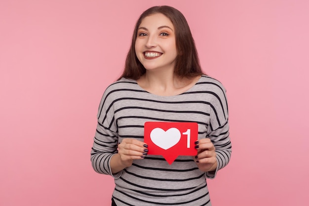 Click Like and follow my blog. Portrait of happy blogger woman in striped sweatshirt holding social media Heart button and looking at camera with toothy smile. studio shot isolated on pink background