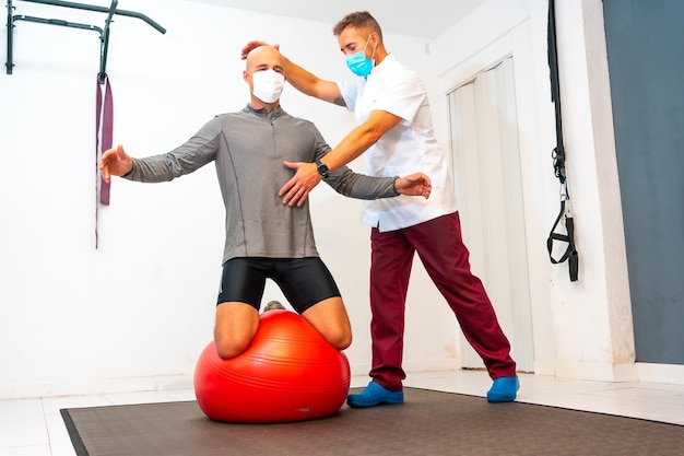 Cliente in equilibrio su pallone rosso assistito da fisioterapista con mascherina fisioterapia con misure protettive per la pandemia di coronavirus covid19 osteopatia quiromassaggio sportivo