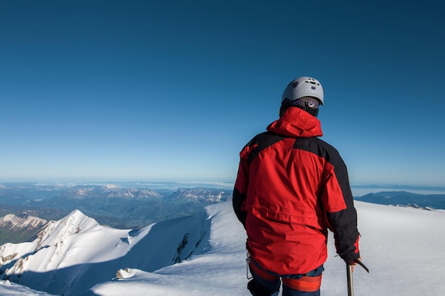 Альпинист в красной куртке стоит на вершине горы на фоне голубого небаxD