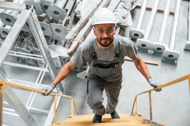 Foto si arrampica sulla scala giovane operaio di fabbrica in uniforme grigio