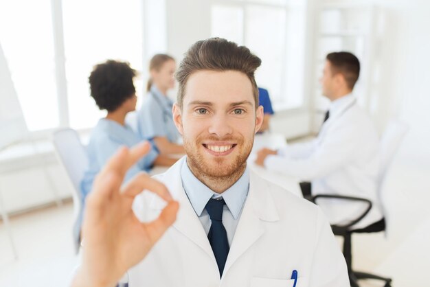 clinic, profession, gesture, people and medicine concept - happy male doctor showing ok hand sign over group of medics meeting at hospital