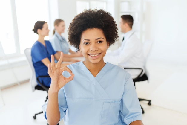 клиника, профессия, люди и концепция медицины - счастливая афро-американская женщина-врач над группой медиков, встречающихся в больнице, показывая знак рукой ок