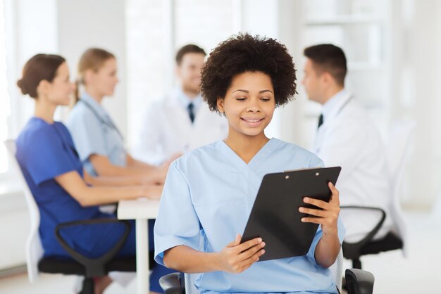 clinic, profession, people and medicine concept - happy female doctor or nurse with clipboard over group of medics meeting at hospital