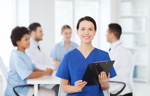 Clinic, profession, people and medicine concept - happy female\
doctor or nurse with clipboard over group of medics meeting at\
hospital