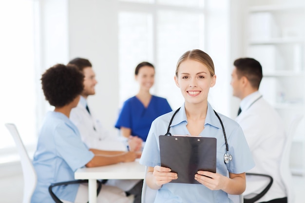 Clinic, profession, people and medicine concept - happy female\
doctor with clipboard over group of medics meeting at hospital