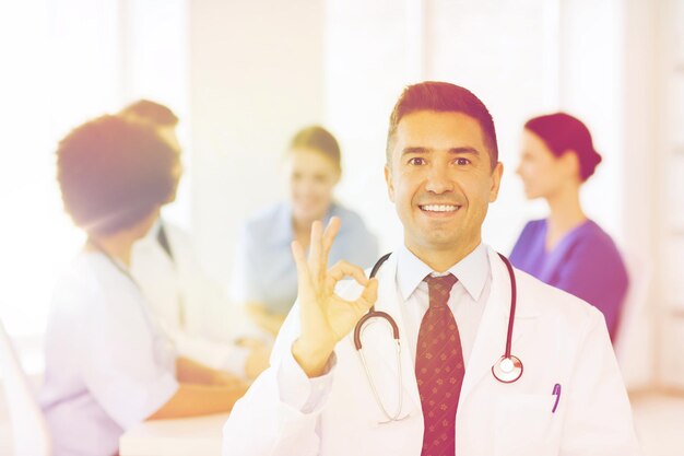 Photo clinic, profession, people and medicine concept - happy male doctor over group of medics meeting at hospital showing ok hand sign