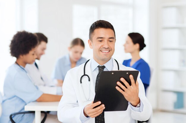 clinic, profession, people and medicine concept - happy male doctor with clipboard over group of medics meeting at hospital