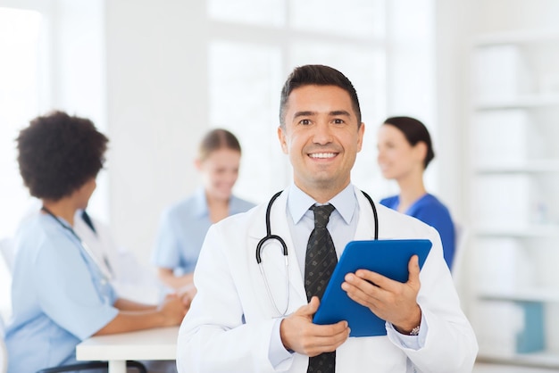 Clinic, profession, people and medicine concept - happy male\
doctor with tablet pc computer over group of medics meeting at\
hospital