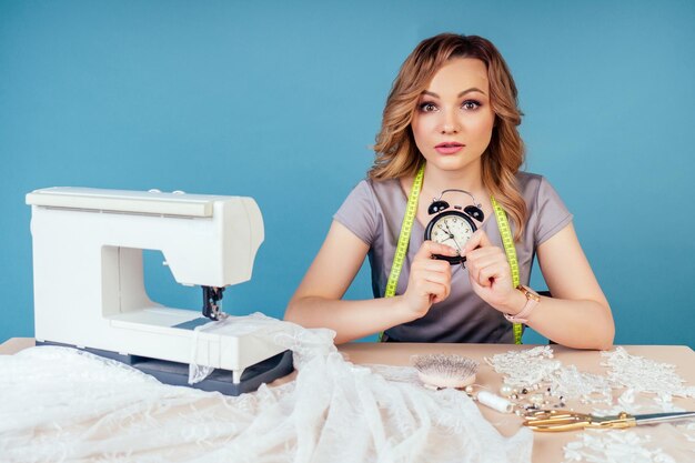 Clock closeup on the table behind woman seamstress tailor dressmaker designer sews a wedding dress with sewing machine on a blue background in the studio concept of deadline