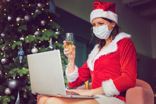 Closdup beautiful asian woman hat and red Santa Claus smiling happily, champagne glass, concept holiday celebrating, Christmas tree background. Christmas greetings online. New year's quarantine.