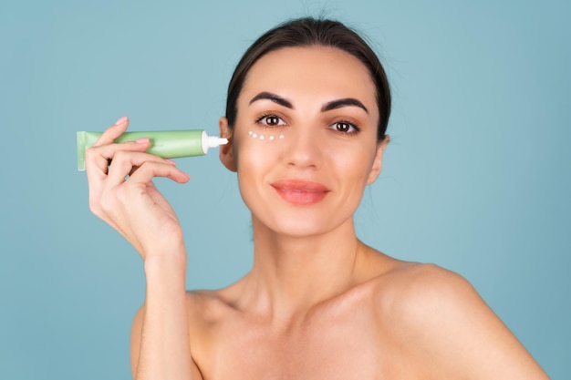 Close beauty portrait of a topless woman with perfect skin and natural make-up, holding a cream for moisturizing and elasticity of the skin under the eyes