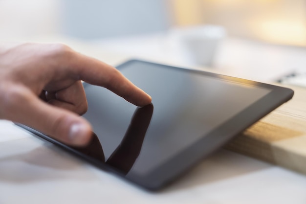 Foto dettaglio ravvicinato del dito di un uomo che entra in contatto con lo schermo di un elegante tablet digitale appoggiato su una scrivania sullo sfondo sfocato di un ufficio pieno di luce
