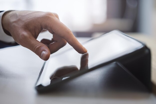 Foto dettaglio ravvicinato del dito di un uomo che entra in contatto con lo schermo di un elegante tablet digitale appoggiato su una scrivania sullo sfondo sfocato di un ufficio pieno di luce