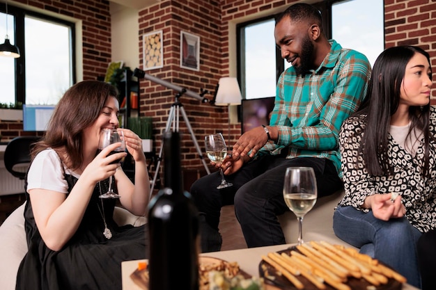 집에 있는 거실에서 유대감을 형성하면서 와인 파티에서 즐거운 시간을 보내는 다양한 친구들과 친해집니다. 함께 시간을 즐기면서 알코올 음료와 스낵으로 생일 이벤트를 축하하는 쾌활한 사람들.