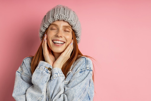 Close eyes. Lovely female with healthy skin, red hair dressed in denim jacket, bulky knitted hat with dreamy expression aside, remembering pleasant moments, isolated over pink background.