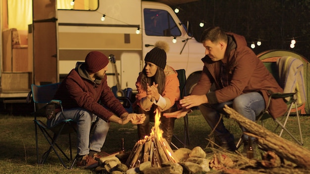 Close friend sitting on camping chairs around camp fire warming their hands. Retro camper. Light bulbs in the background.