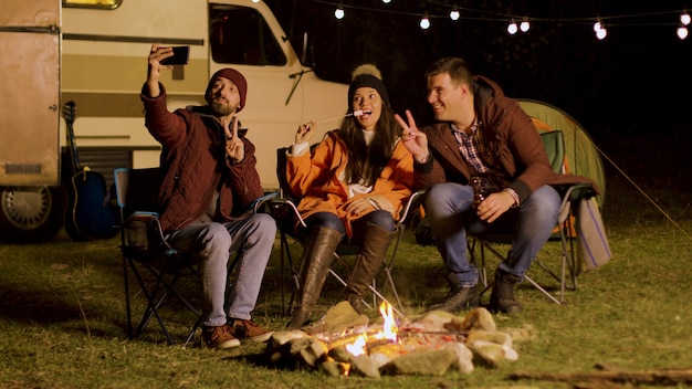 Close friends taking a selfie around camp fire in a cold night of autumn in the mountains. Retro camper van. Light bulbs in the background.
