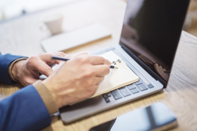 Foto prospettiva ravvicinata della mano di un uomo che inserisce voci in un blocco note su un laptop all'avanguardia con una morbida sfocatura nell'ambiente circostante