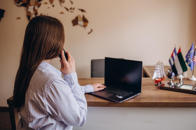 Close photo of female travel agent young woman talking on phone
smiling and looking at camera travel agency office interior with
big world map