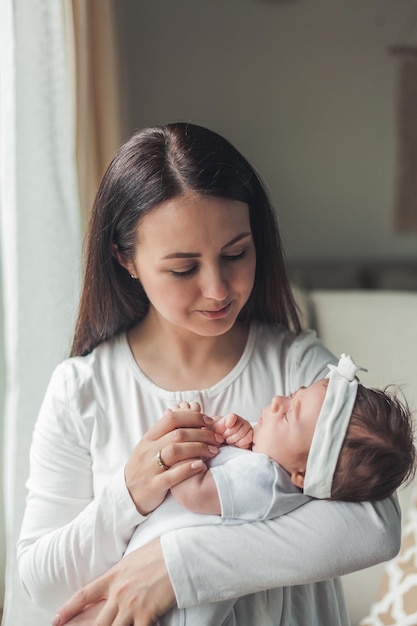 Ritratto ravvicinato di una piccola neonata carina dai capelli scuri in un abito bianco tra le braccia di una giovane madre genitore di maternità sonno sano