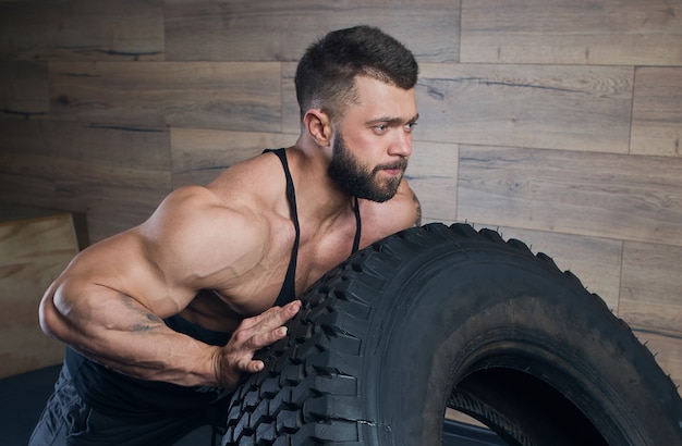 Close portrait of strong man in black tank top and space gray shorts with a beard who tries to push a tire in gym