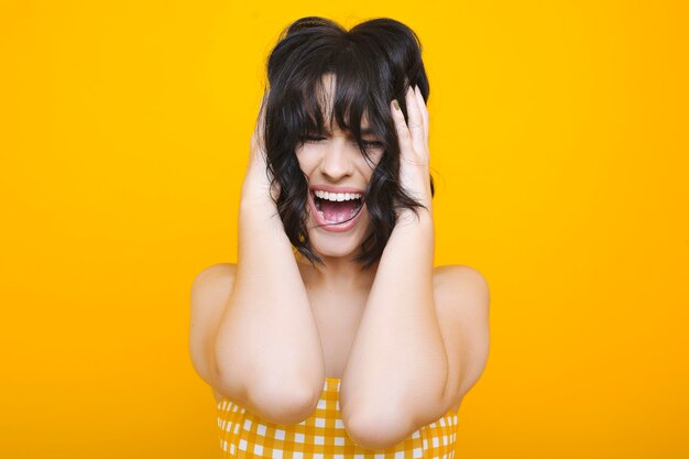 Close up of a adult caucasian female with short dark hair screaming with closed eyes while holding hair with both hands in front of a yellow wall dressed in yellow