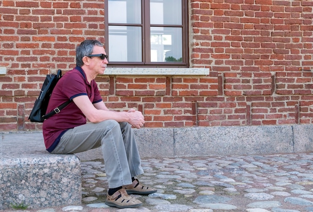 Close-up of an adult pensioner in summer clothes and with a backpack sits and rests against the backdrop of a red brick building. Life style. Selective focus.