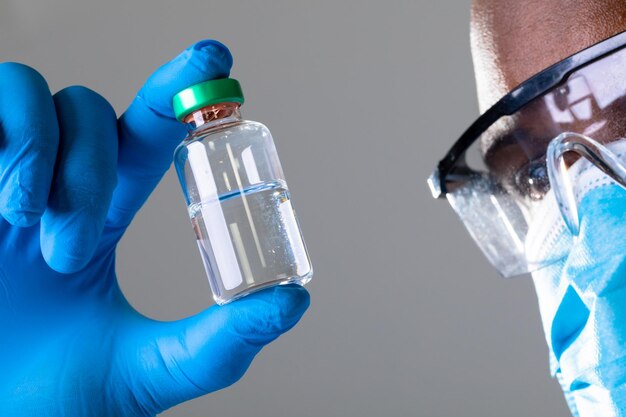 Close up of african american male health worker wearing face mask holding covid-19 vaccine bottle