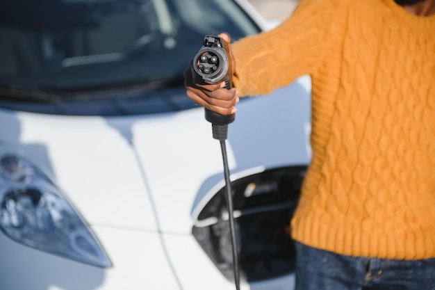 Close up of african american man connecting charging cable to\
electric car. young male standing near his modern auto with leather\
suitcase in hand.