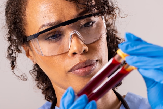 Foto primo piano del medico femminile di metà adulto afroamericano che indossa occhiali protettivi tenendo la provetta. concetto di sangue, provetta, medicina, campione, laboratorio, medico, assistenza sanitaria e occupazione medica.