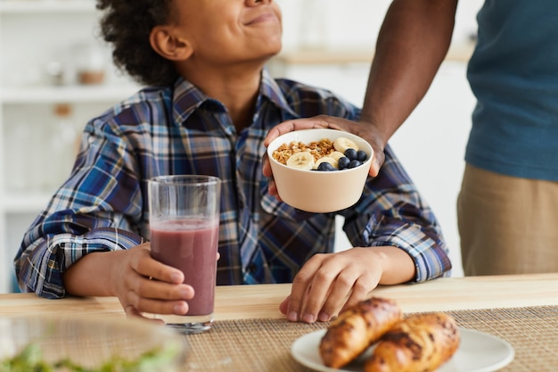 Primo piano del ragazzo africano che mangia cereali e che beve succo di frutta fresco per colazione preparata da suo padre