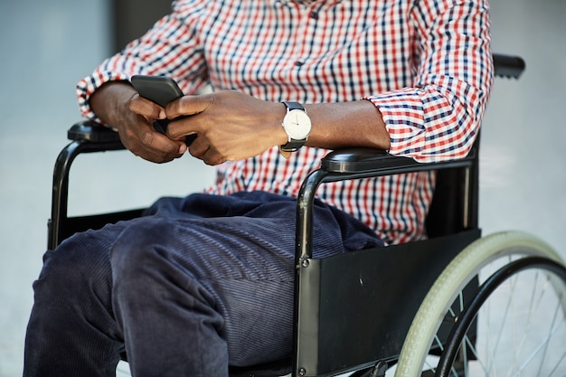 Primo piano dell'uomo africano disabile che si siede in sedia a rotelle e che gioca sul suo telefono cellulare