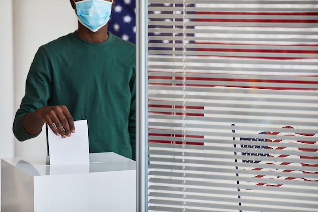 Photo close-up of afro-american man in mask putting the ballot into the box at polling station during pandemic