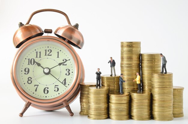 Close-up of alarm clock by figurines on coins stack against white background