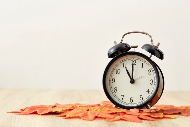 Photo close-up of alarm clock with autumn leaves on table against wall