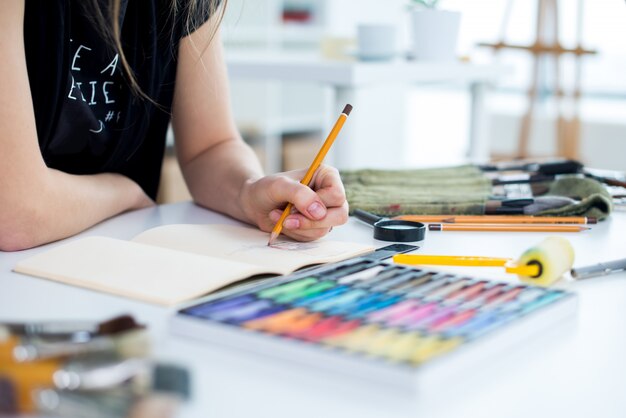 Close-up angle view of a female painter drawing draft at sketchbook using pencil. Artist sketching in art studio with crayons set.