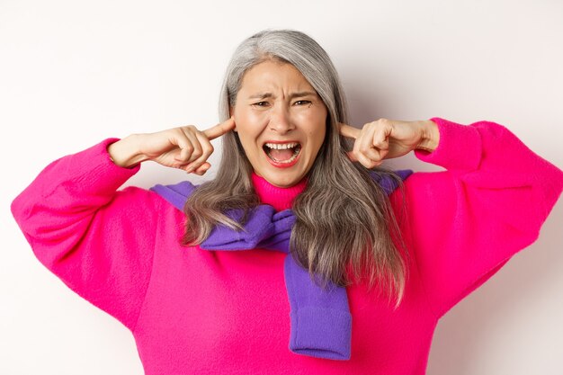 Close-up of annoyed and bothered asian senior woman screaming with shut ears, disturbed by loud noise, standing on white.