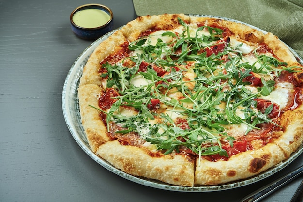 Close up appetizing pizza with prosciutto, arugula, tomatoes, parmesan and mozzarella on wooden table in a restaurant. Selective focus, copy space