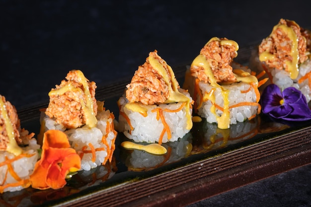 Close up of appetizing sushi topped with salmon and fresh sauce served on wooden tray with flowers in restaurant against black background