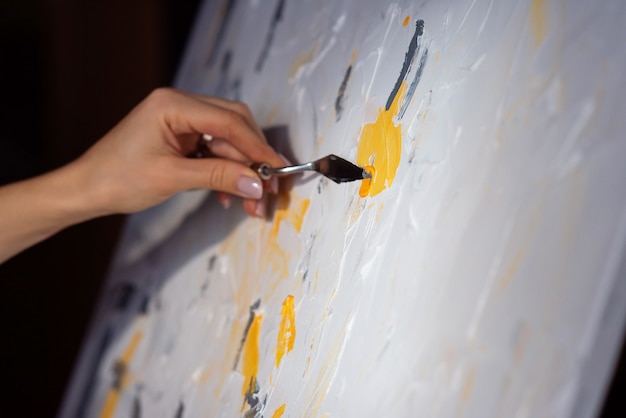 Close up artist's hand with mastic and yellow paint slides on white canvas. Art and creative concept.