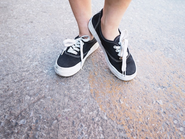コンクリートの道に立っている黒いスニーカーでアジアの女の子の足を閉じます。流行に敏感なライフスタイル。