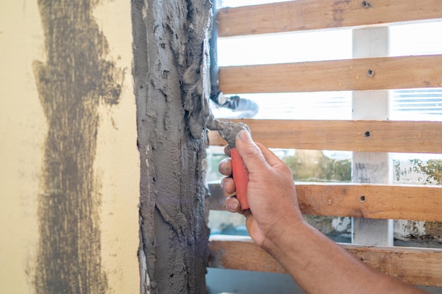 Primo piano la mano dell'uomo asiatico tiene la cazzuola per riparare e decorare la parete con cemento fresco con stecche di legno accanto