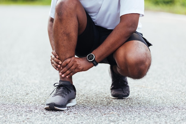 Close up Asian young sport runner black man wear watch hands joint hold leg pain because of twisted ankle broken while running at the outdoor street health park, healthy exercise Injury from workout