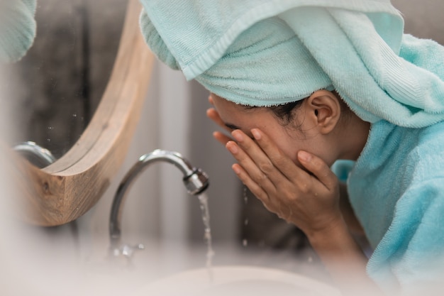 Foto chiuda in su della giovane donna asiatica con un asciugamano sulla sua testa in piedi in bagno, lavandosi il viso