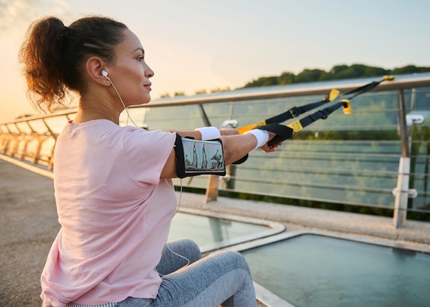 Close-up athletic sportswoman doing exercise with straps during intense suspension training at the urban environment in summer.Female athlete in sportswear pulling suspension ropes, squatting, lunging