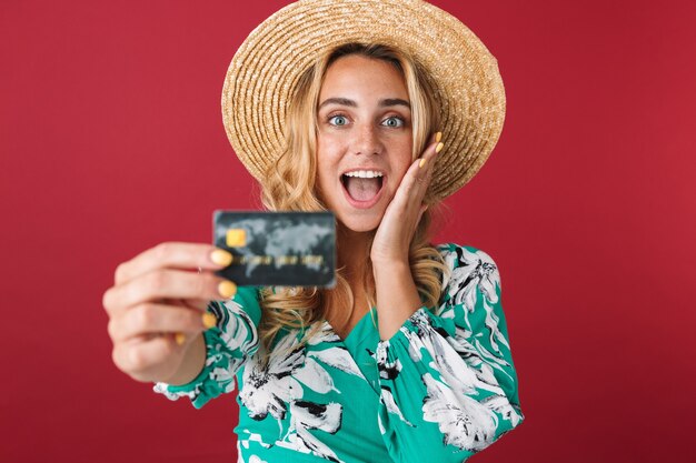 Close up of an attractive excited young blonde woman wearing summer dress and straw hat standing isolated over pink wall, showing credit card